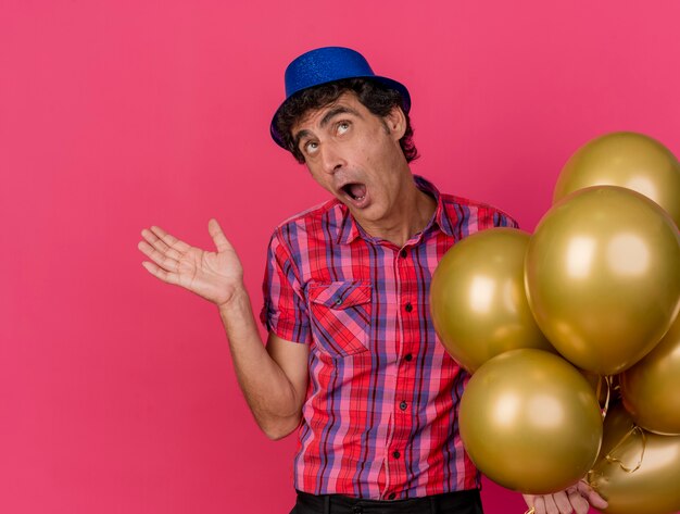 Impressionné homme de fête d'âge moyen portant chapeau de fête tenant des ballons à la recherche montrant la main vide isolé sur mur cramoisi