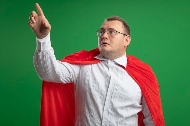 Impressionné homme de super-héros slaves adultes en cape rouge portant des lunettes à la recherche et pointant vers le haut isolé sur fond vert