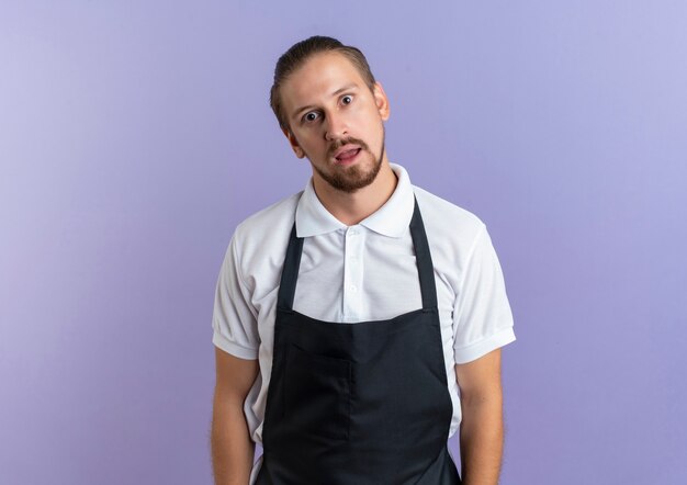 Photo gratuite impressionné jeune beau coiffeur en uniforme debout et à la isolé sur violet avec copie espace