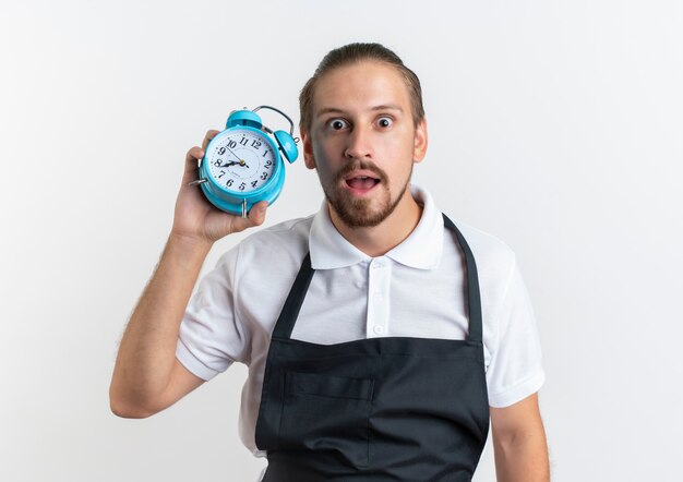 Impressionné jeune beau coiffeur en uniforme tenant un réveil isolé sur blanc