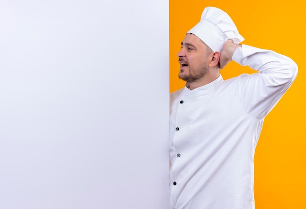 Impressionné jeune beau cuisinier en uniforme de chef debout derrière un mur blanc pointant la main dessus isolé sur un mur orange avec espace de copie