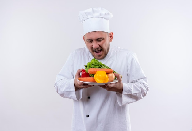 Impressionné jeune beau cuisinier en uniforme de chef tenant une assiette avec des légumes en les regardant sur un mur blanc isolé