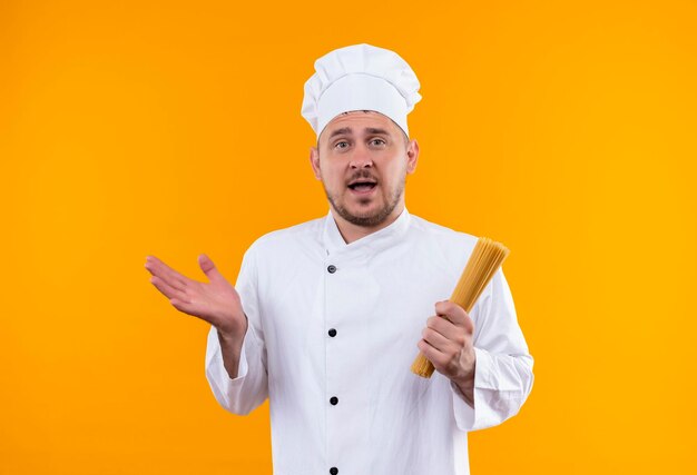 Impressionné jeune beau cuisinier en uniforme de chef tenant des pâtes spaghetti et montrant une main vide isolée sur un mur orange