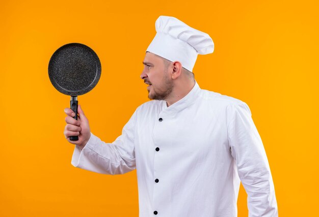 Impressionné jeune beau cuisinier en uniforme de chef tenant une poêle à frire regardant le côté isolé sur un mur orange