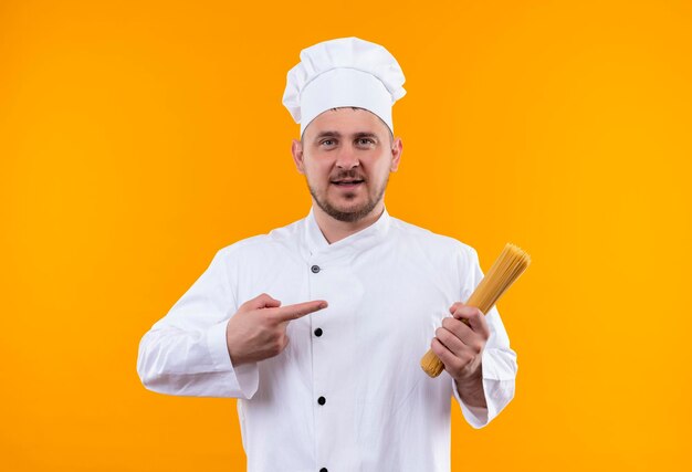 Impressionné jeune beau cuisinier en uniforme de chef tenant et pointant sur des pâtes spaghetti isolées sur un mur orange