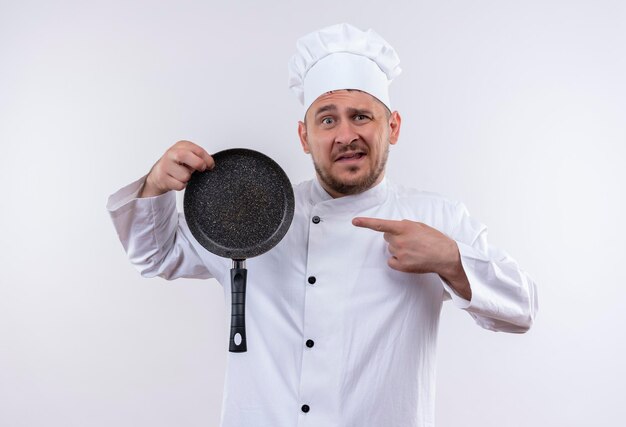 Impressionné jeune beau cuisinier en uniforme de chef tenant et pointant sur une poêle à frire isolée sur un mur blanc