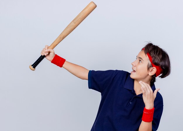Impressionné jeune beau garçon sportif portant un bandeau et des bracelets avec des appareils dentaires soulevant une batte de baseball en le regardant en gardant la main dans l'air isolé sur fond blanc