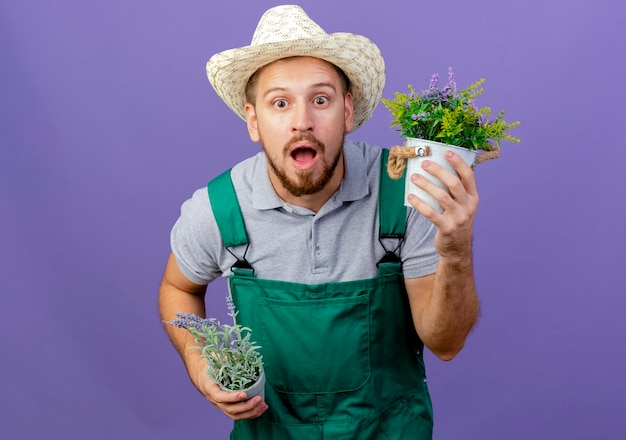 Impressionné jeune beau jardinier slave en uniforme et chapeau tenant des pots de fleurs isolés sur mur violet avec espace de copie