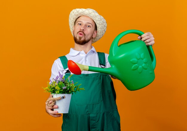 Impressionné jeune beau jardinier slave en uniforme et hat holding pot de fleurs et arrosoir à isolé