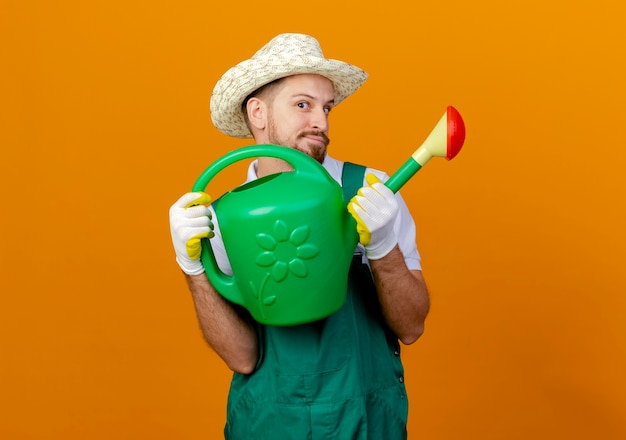 Impressionné jeune beau jardinier slave en uniforme portant un chapeau et des gants de jardinage tenant un arrosoir à la recherche d'isolement