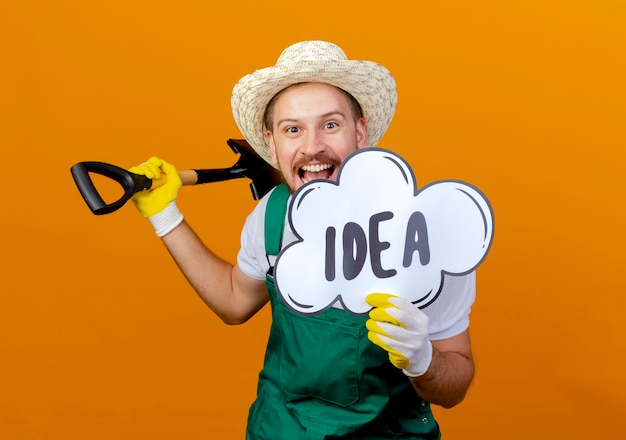 Photo gratuite impressionné jeune beau jardinier slave en uniforme portant un chapeau et des gants de jardinage tenant une bêche et une bulle d'idée à isolé