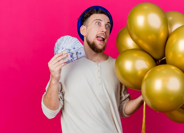 Impressionné jeune beau mec du parti slave portant chapeau de fête tenant des ballons et de l'argent montrant la langue à côté isolé sur mur rose avec espace copie