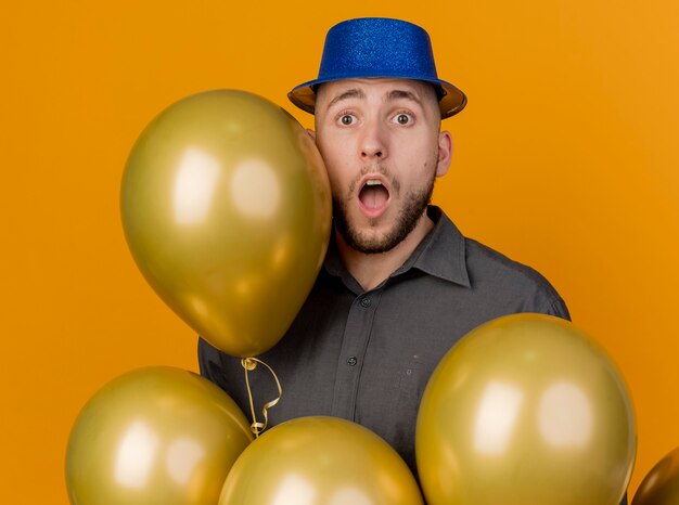 Impressionné jeune beau mec de fête slave portant chapeau de fête debout derrière des ballons regardant la caméra isolée sur fond orange