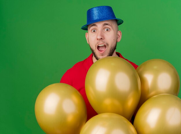 Impressionné jeune beau mec de fête slave portant chapeau de fête debout derrière des ballons regardant la caméra isolée sur fond vert