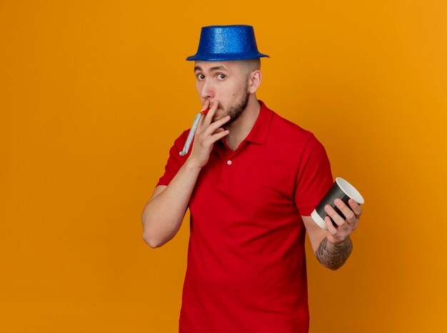 Impressionné jeune beau mec de fête slave portant chapeau de fête regardant la caméra tenant le ventilateur de fête dans la bouche avec une tasse de café en plastique à la main isolé sur fond orange avec espace de copie