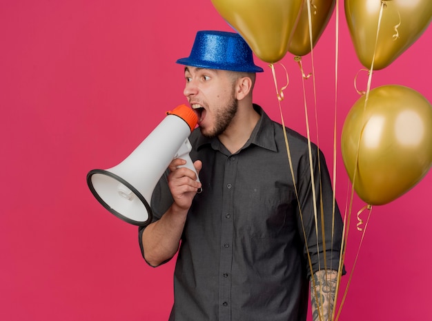 Impressionné jeune beau mec de fête slave portant chapeau de fête tenant des ballons en criant dans haut-parleur à côté isolé sur fond cramoisi