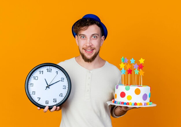 Impressionné jeune beau mec de fête slave portant chapeau de fête tenant horloge et gâteau d'anniversaire avec des étoiles regardant la caméra isolée sur fond orange