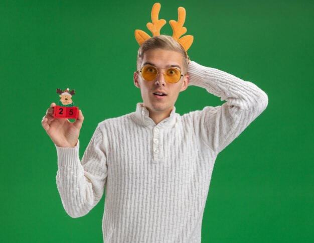 Impressionné jeune beau mec portant bandeau de bois de renne avec des lunettes tenant jouet arbre de Noël avec date regardant la caméra en gardant la main derrière la tête isolé sur fond vert