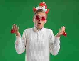 Photo gratuite impressionné jeune beau mec portant bandeau du père noël avec des lunettes tenant des boules d'ornement de noël isolé sur mur vert