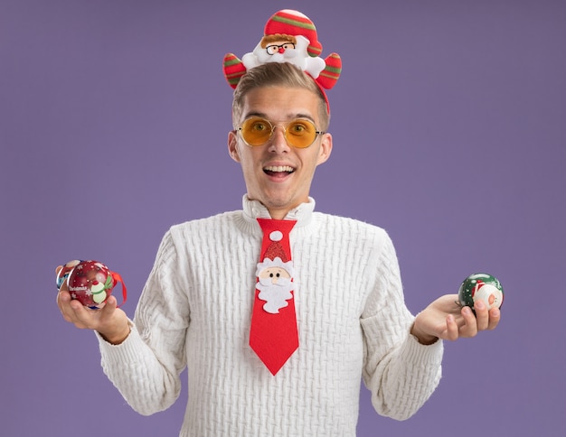 Photo gratuite impressionné jeune beau mec portant bandeau de père noël et cravate avec des lunettes tenant des ornements de boule de noël regardant la caméra isolée sur fond violet