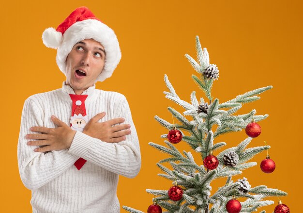 Impressionné jeune beau mec portant chapeau de Noël et cravate de père Noël debout près de sapin de Noël décoré en gardant les mains croisées sur la poitrine à côté isolé sur fond orange