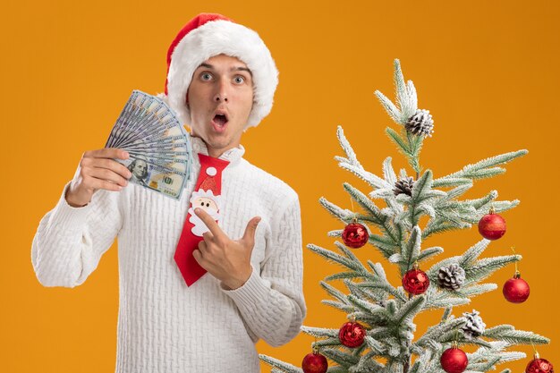 Impressionné jeune beau mec portant chapeau de Noël et cravate de père Noël debout près de sapin de Noël décoré tenant et pointant vers l'argent en regardant la caméra isolée sur fond orange