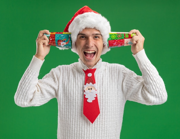 Photo gratuite impressionné jeune beau mec portant chapeau de noël et cravate de père noël tenant des tasses de noël en plastique à côté des oreilles isolé sur mur vert