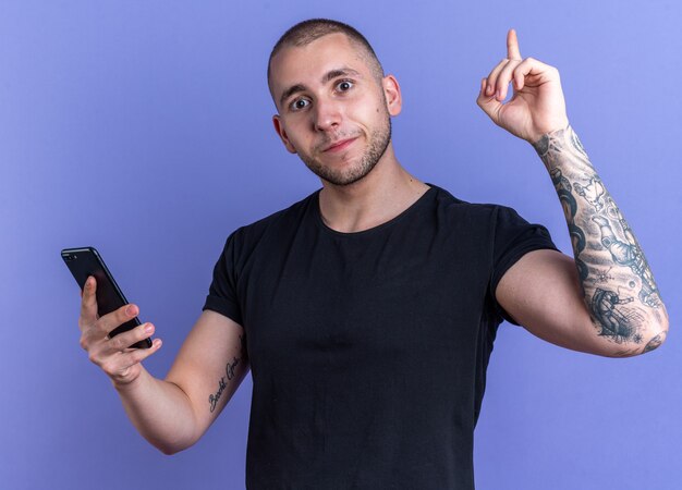 Impressionné jeune beau mec portant un t-shirt noir tenant des points de téléphone isolés sur un mur bleu