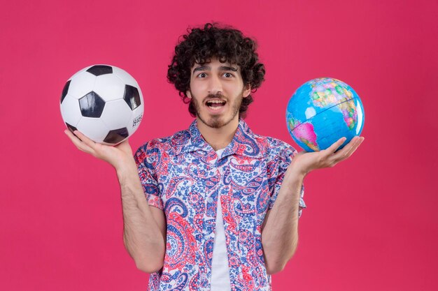 Impressionné jeune beau voyageur bouclé homme tenant un globe et un ballon de football sur un espace rose isolé
