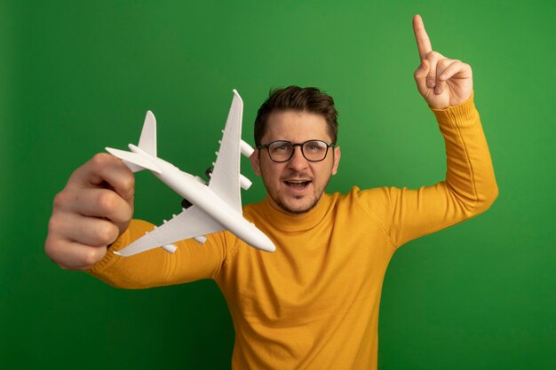 Impressionné jeune bel homme blond portant des lunettes étirant l'avion modèle pointant vers le haut à isolé sur mur vert