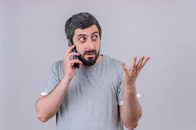 Impressionné jeune bel homme caucasien parlant au téléphone en regardant sur le côté et montrant la main vide isolé sur blanc avec espace copie