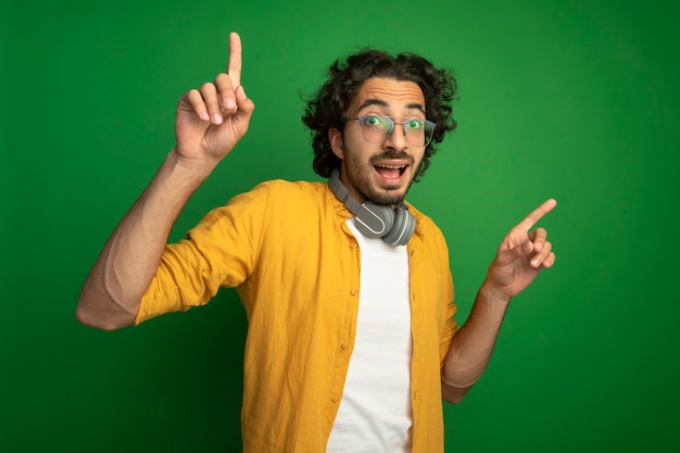 Impressionné Jeune Bel Homme Caucasien Portant Des Lunettes Avec Des écouteurs Autour Du Cou Regardant La Caméra Vers Le Haut Isolé Sur Fond Vert