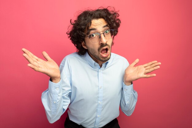 Impressionné jeune bel homme caucasien portant des lunettes regardant la caméra montrant les mains vides isolés sur fond cramoisi