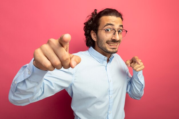 Impressionné jeune bel homme caucasien portant des lunettes regardant la caméra en souriant et en vous faisant geste isolé sur fond cramoisi