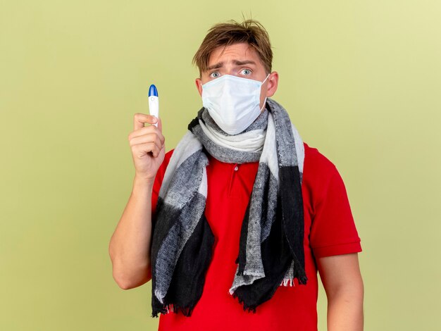 Impressionné jeune bel homme malade blonde portant un masque et une écharpe tenant un thermomètre isolé sur un mur vert olive avec espace copie