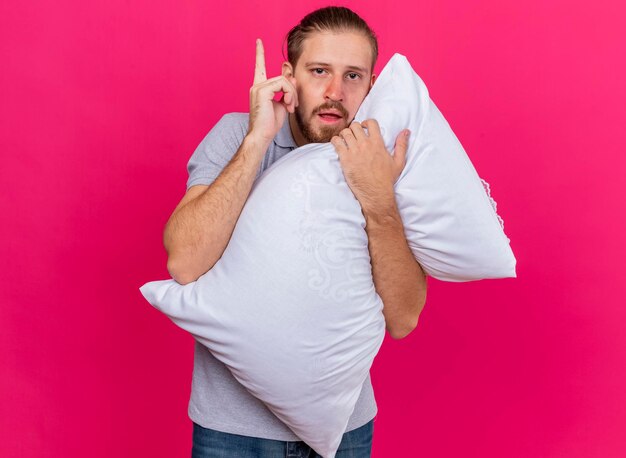 Impressionné jeune bel homme malade slave hugging oreiller à l'avant levant le doigt isolé sur mur rose avec copie espace