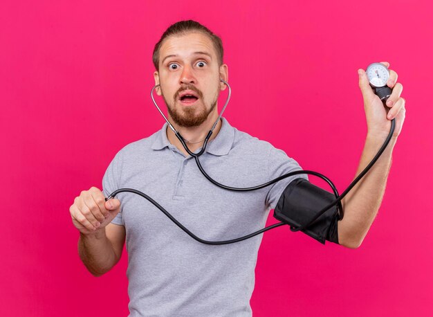 Impressionné jeune bel homme malade slave portant un stéthoscope mesurant sa pression avec un sphygmomanomètre à l'avant isolé sur un mur rose