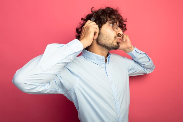 Impressionné jeune bel homme portant des lunettes et des écouteurs saisissant des écouteurs faisant le geste de baiser à côté isolé sur le mur rose