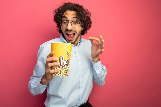 Impressionné jeune bel homme portant des lunettes tenant et regardant à l'intérieur du seau de pop-corn et tenant le morceau de pop-corn isolé sur mur rose