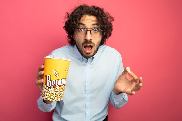 Impressionné jeune bel homme portant des lunettes tenant un seau de pop-corn et morceau de pop-corn à l'avant isolé sur mur rose