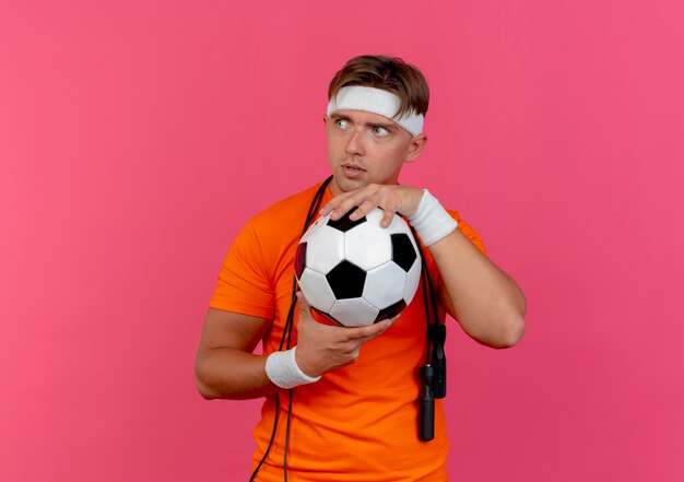 Impressionné jeune bel homme sportif portant bandeau et bracelets avec corde à sauter autour du cou tenant un ballon de soccer à côté isolé sur rose