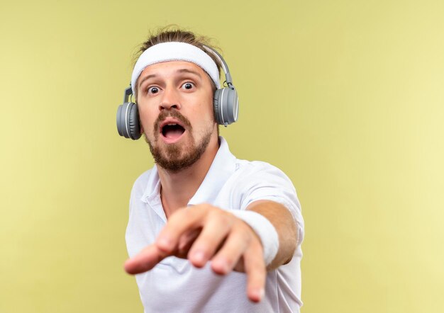 Impressionné jeune bel homme sportif portant un bandeau et des bracelets et des écouteurs tendant la main vers isolé sur un mur vert avec espace de copie