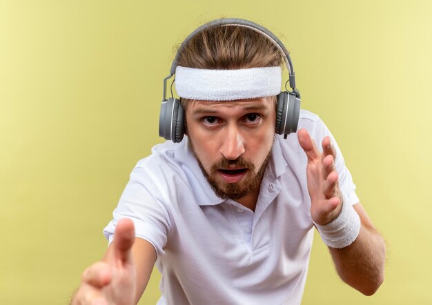 Impressionné jeune bel homme sportif portant un bandeau et des bracelets et des écouteurs tendant les mains et isolé sur un mur vert