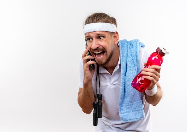 Impressionné jeune bel homme sportif portant un bandeau et des bracelets parlant au téléphone tenant une bouteille d'eau avec une serviette et une corde à sauter sur les épaules isolées sur un mur blanc avec espace de copie