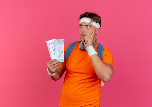 Impressionné jeune bel homme sportif portant un bandeau et des bracelets et un sac à dos avec des écouteurs sur le cou tenant des billets d'avion mettant la main sur le visage en regardant de côté