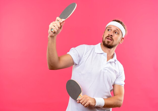 Impressionné jeune bel homme sportif portant un bandeau et des bracelets tenant des raquettes de ping-pong et les regardant isolés sur un mur rose avec espace de copie