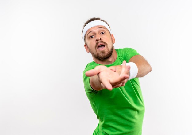 Impressionné jeune bel homme sportif portant un bandeau et des bracelets tendant la main isolée sur un mur blanc avec un espace de copie