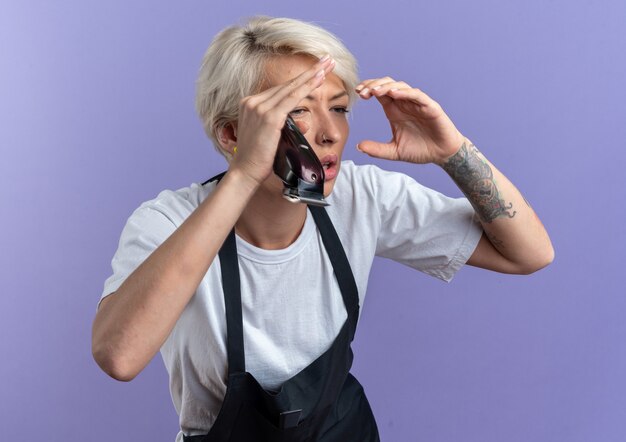 Impressionné jeune belle femme barbier en uniforme tenant une tondeuse à cheveux à la distance avec les mains isolées sur le mur bleu