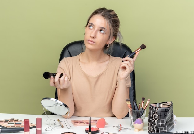 Impressionné jeune belle fille assise au bureau avec des outils de maquillage tenant un pinceau de maquillage isolé sur un mur vert olive