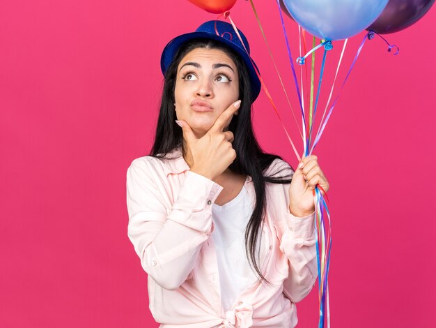 Impressionné jeune belle fille portant un chapeau de fête tenant des ballons attrapé le menton